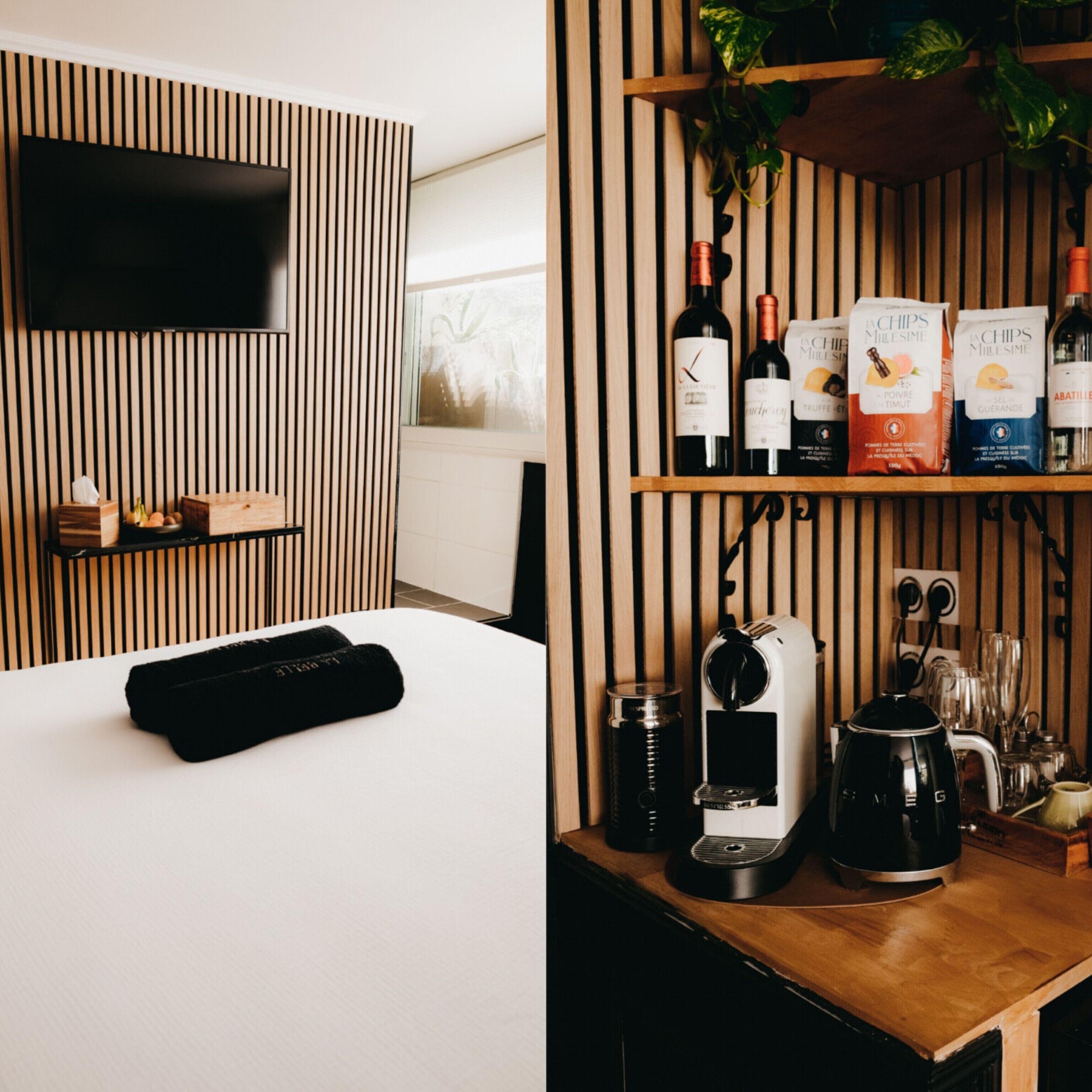 Une chambre d’hôtel moderne avec un mur en lambris de bois, un téléviseur à écran plat, un lit, une machine à café et une étagère contenant des bouteilles de vin et des en-cas.