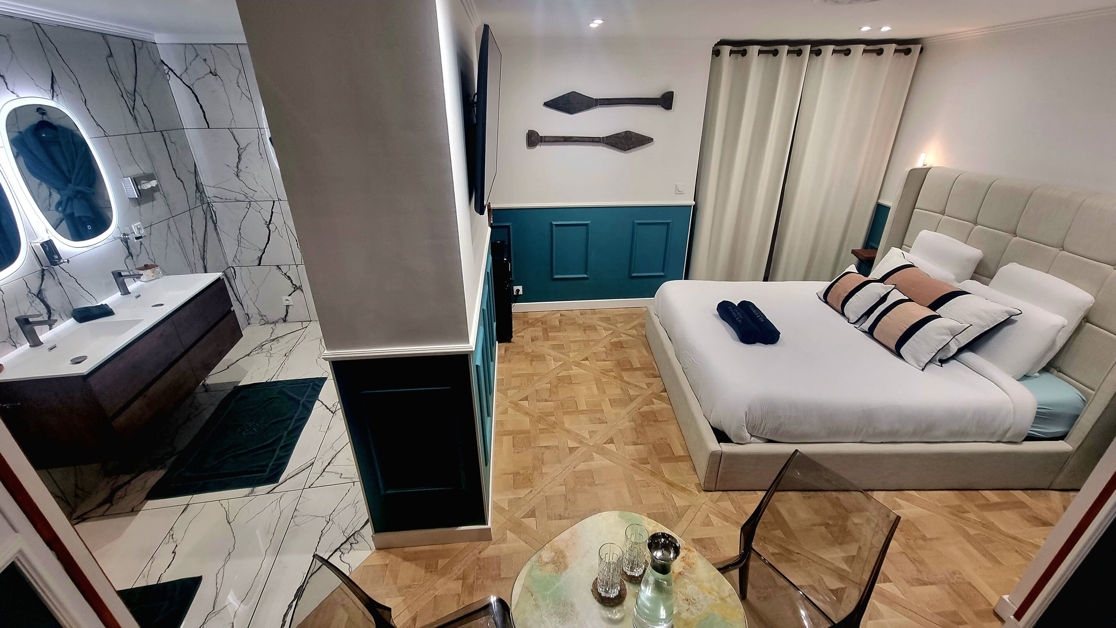 Habitación de hotel con baño de mármol, cama con almohadas, mesita de noche con vasos y televisor en la pared.