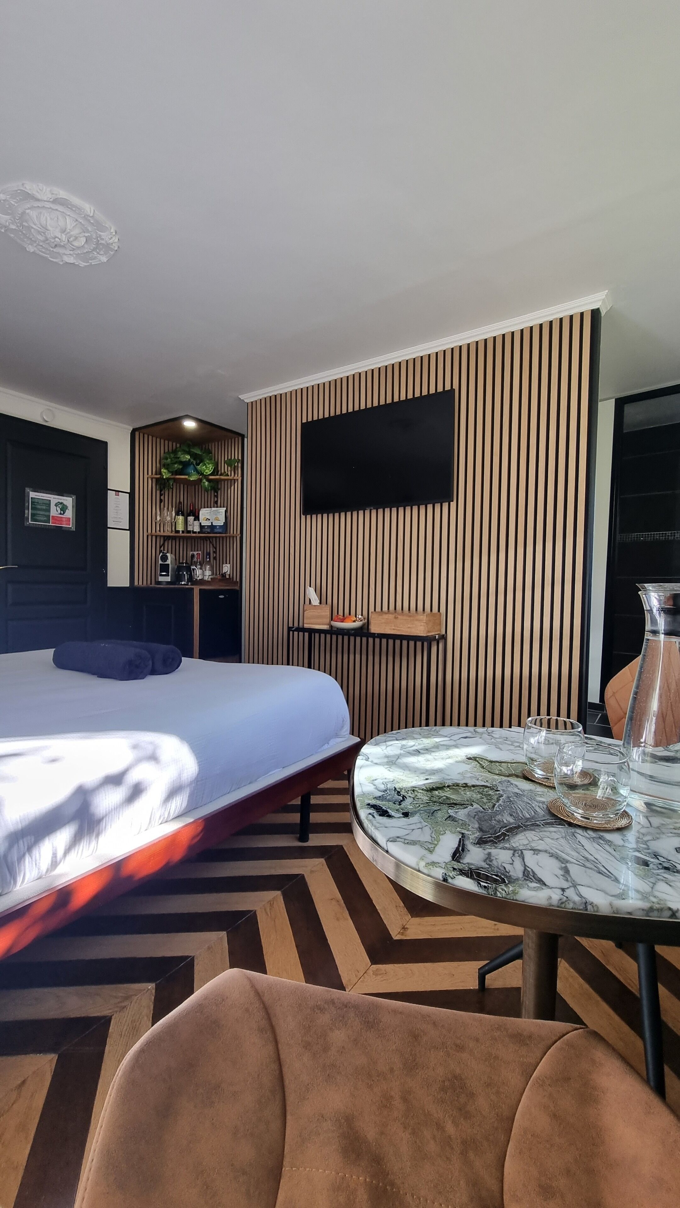Una habitación de hotel moderna con una cama grande, una mesa redonda de mármol, un televisor montado en una pared de paneles de madera, una pequeña kitchenette y una planta en maceta.