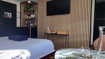 Una habitación de hotel moderna con una cama grande, una mesa redonda de mármol, un televisor montado en una pared de paneles de madera, una pequeña kitchenette y una planta en maceta.