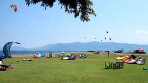 Um campo gramado com vários balões coloridos e tendas, uma praia ao fundo e montanhas ao longe.