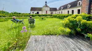 A courtyard with two people relaxing on lawn chairs, a wooden deck, and a historic building with a tower in the background. - Nice little cozy cottage, ideal for a romantic stay (Champlay)
