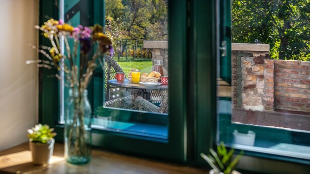 Vista desde una ventana a un patio con una mesa dispuesta para el desayuno, un jarrón con flores y plantas en macetas en el alféizar.