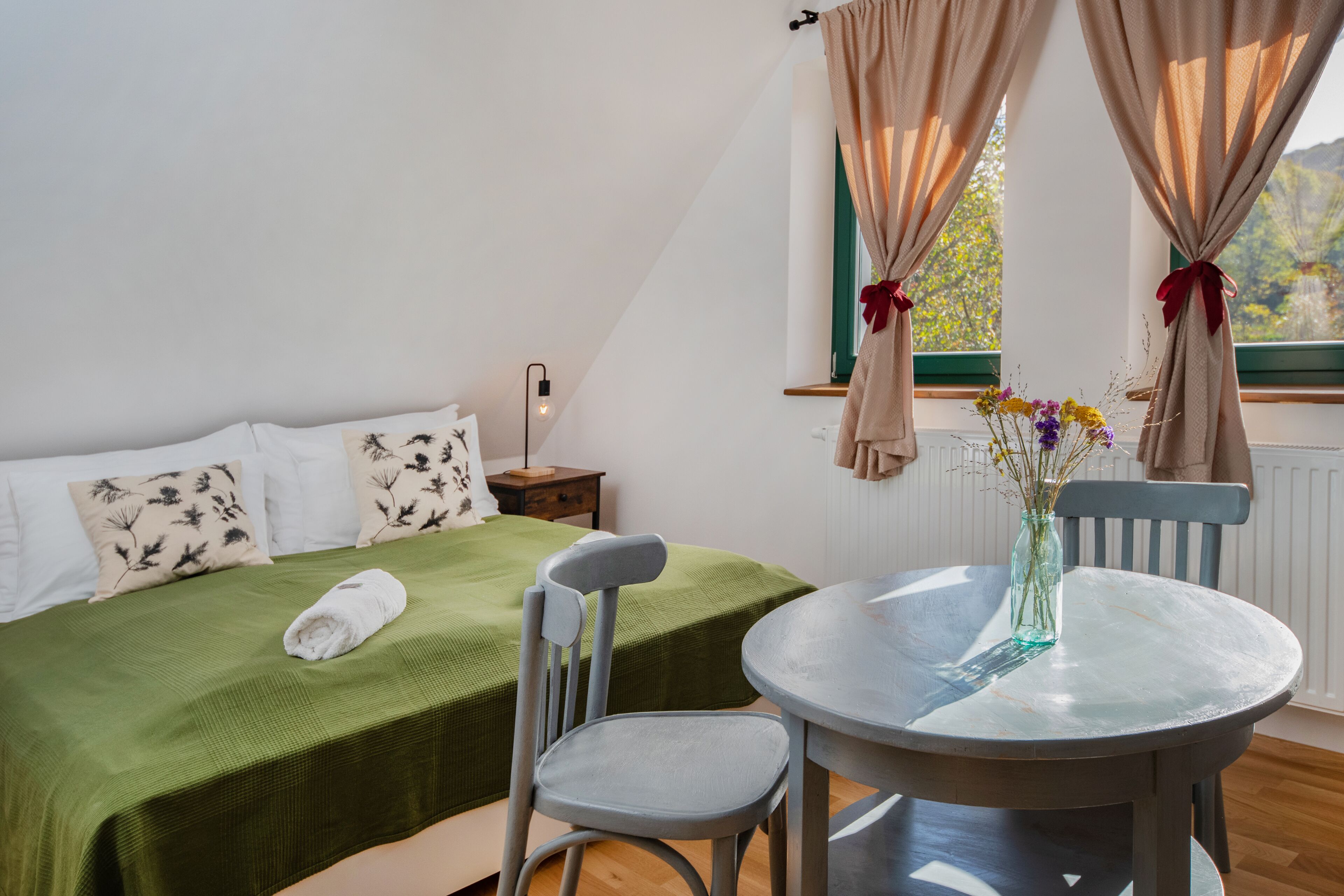 A bedroom with a green bedspread, a neatly folded white towel, a small table with a vase of flowers, and a chair.
