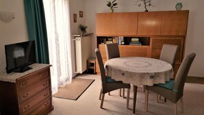 A dining area with a round table, chairs, a television on a dresser, a radiator, and a bookshelf with books and decorative items. - Convenient (Bad Vilbel)