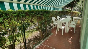 Un espace patio aménagé avec une table et des chaises, entouré de verdure et surmonté d’un auvent à rayures.