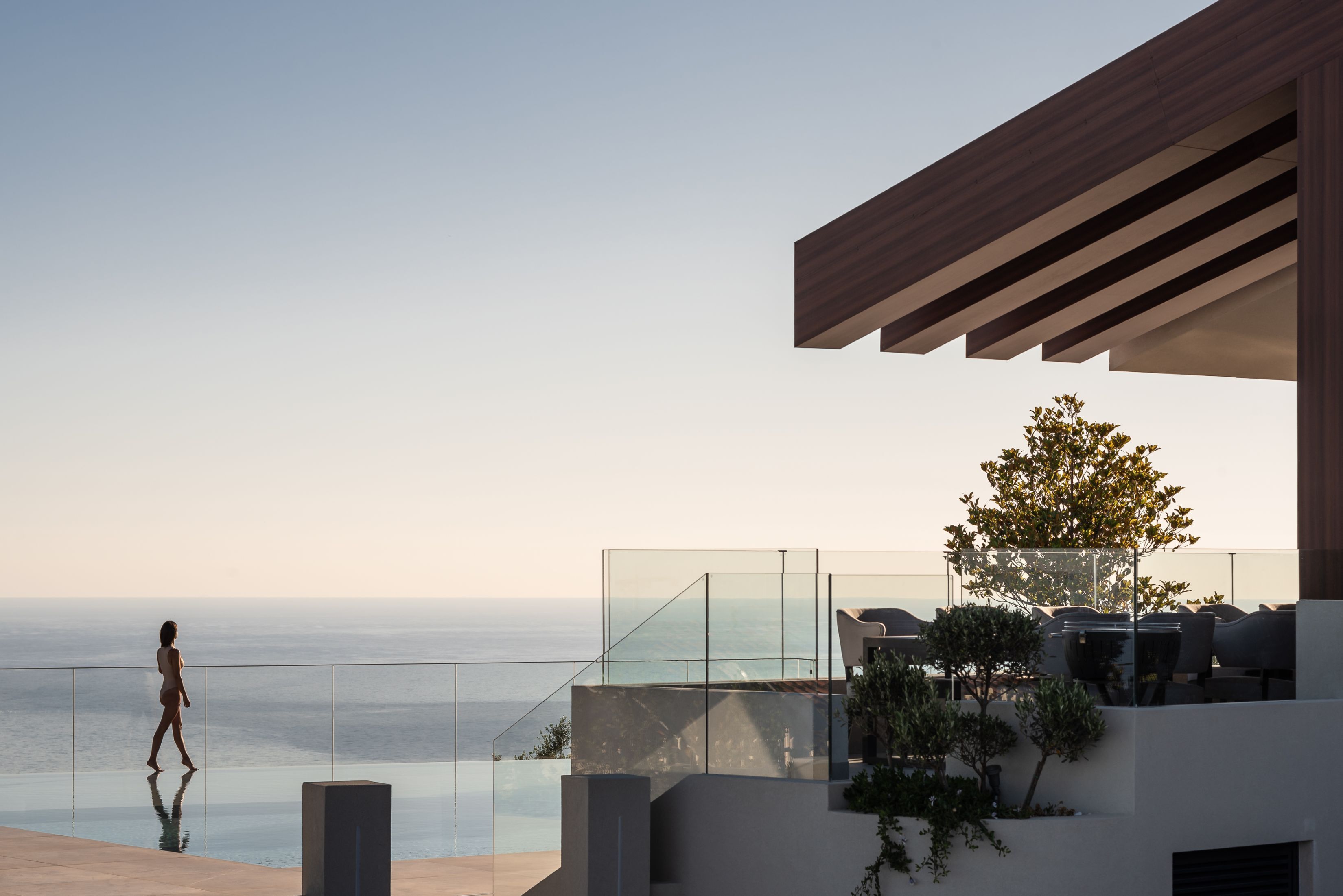 A modern outdoor terrace with a glass balustrade overlooking the sea, a wooden pergola, and a seating area with a tree.