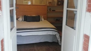 En smal døråbning ind til et soveværelse med en seng, sengegavl, natbord, lampe og en reol med diverse genstande.