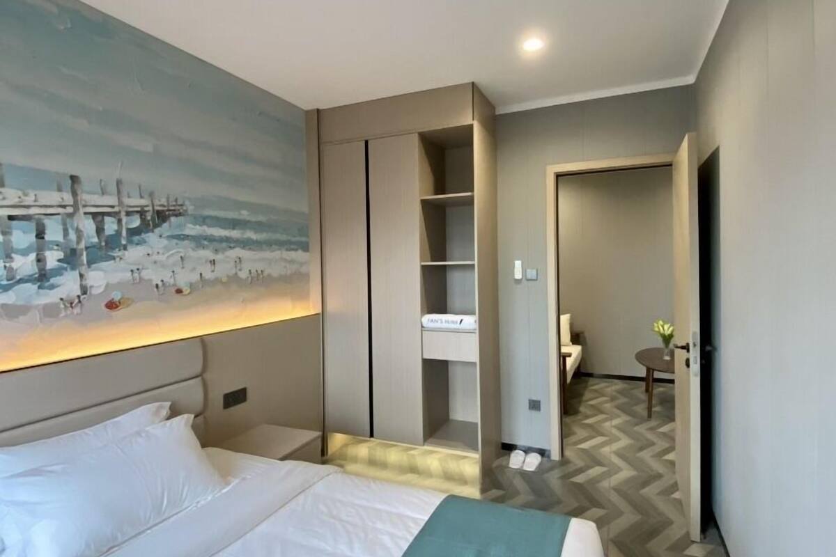 Una habitación de hotel moderna con una cama, un cuadro de un muelle, un armario empotrado y un baño visible a través de una puerta abierta.