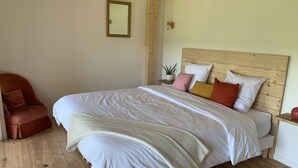 Une chambre avec un grand lit, une tête de lit en bois, une table de chevet, une chaise, une petite plante et un miroir fixé au mur.