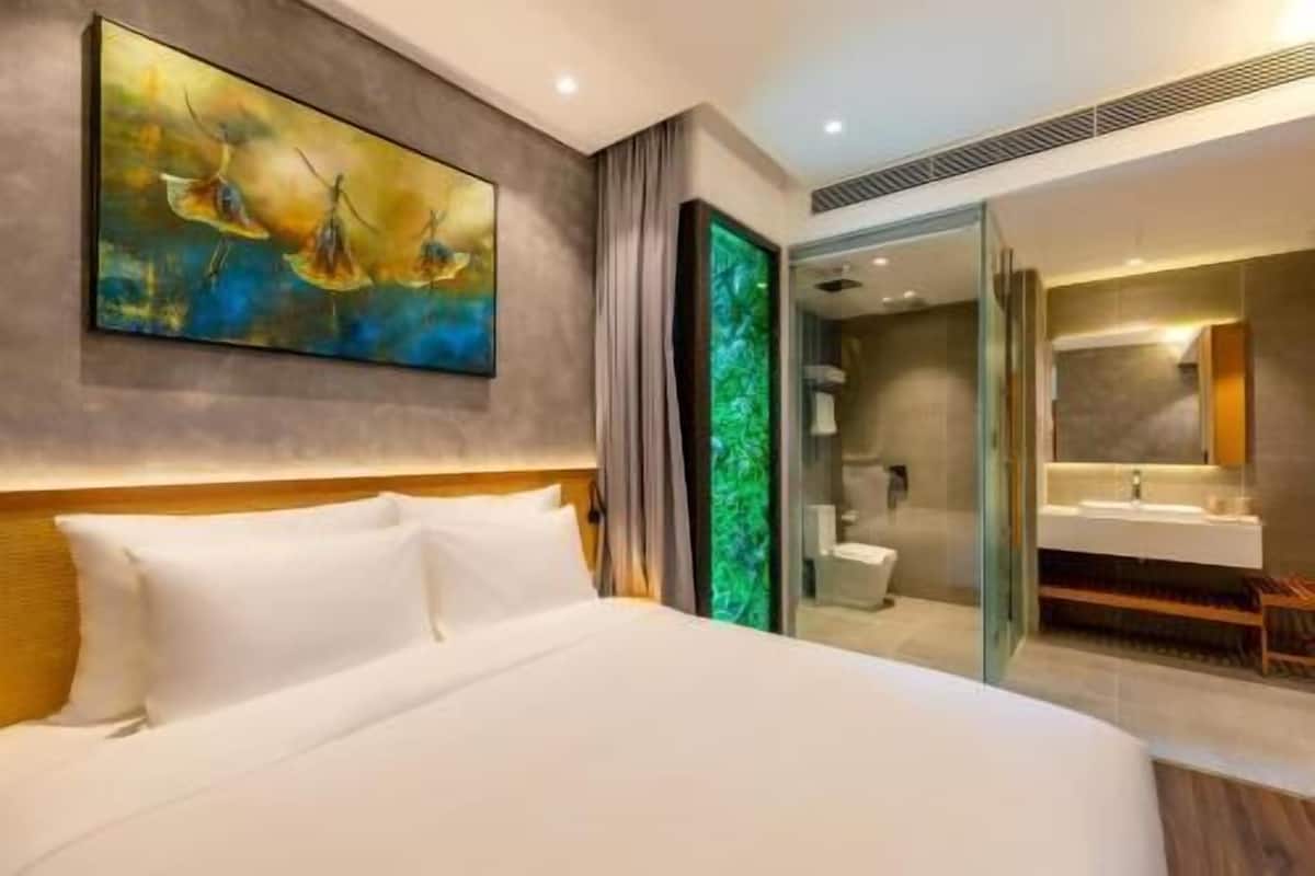 A modern hotel room with a large bed, a painting on the wall, a bathroom with a glass door, and a green accent wall.