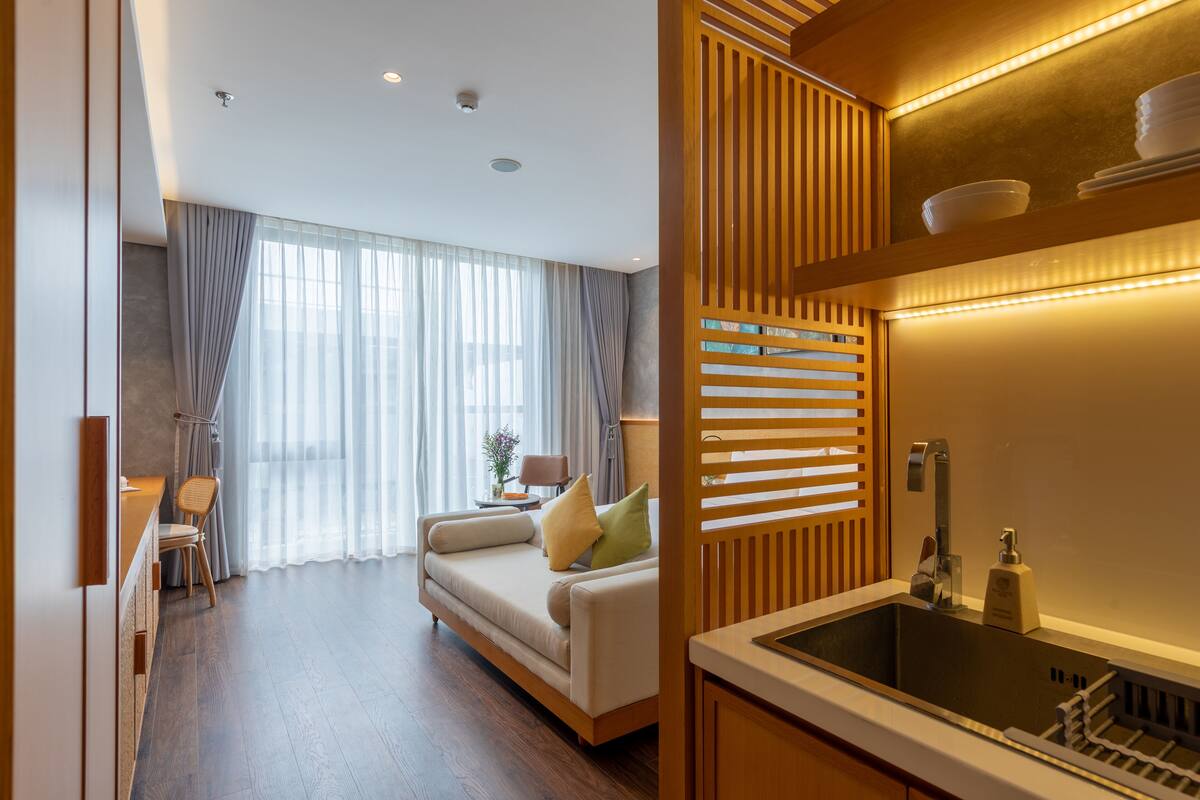 A modern hotel room with a sofa, a dining area, and a kitchen with a sink.