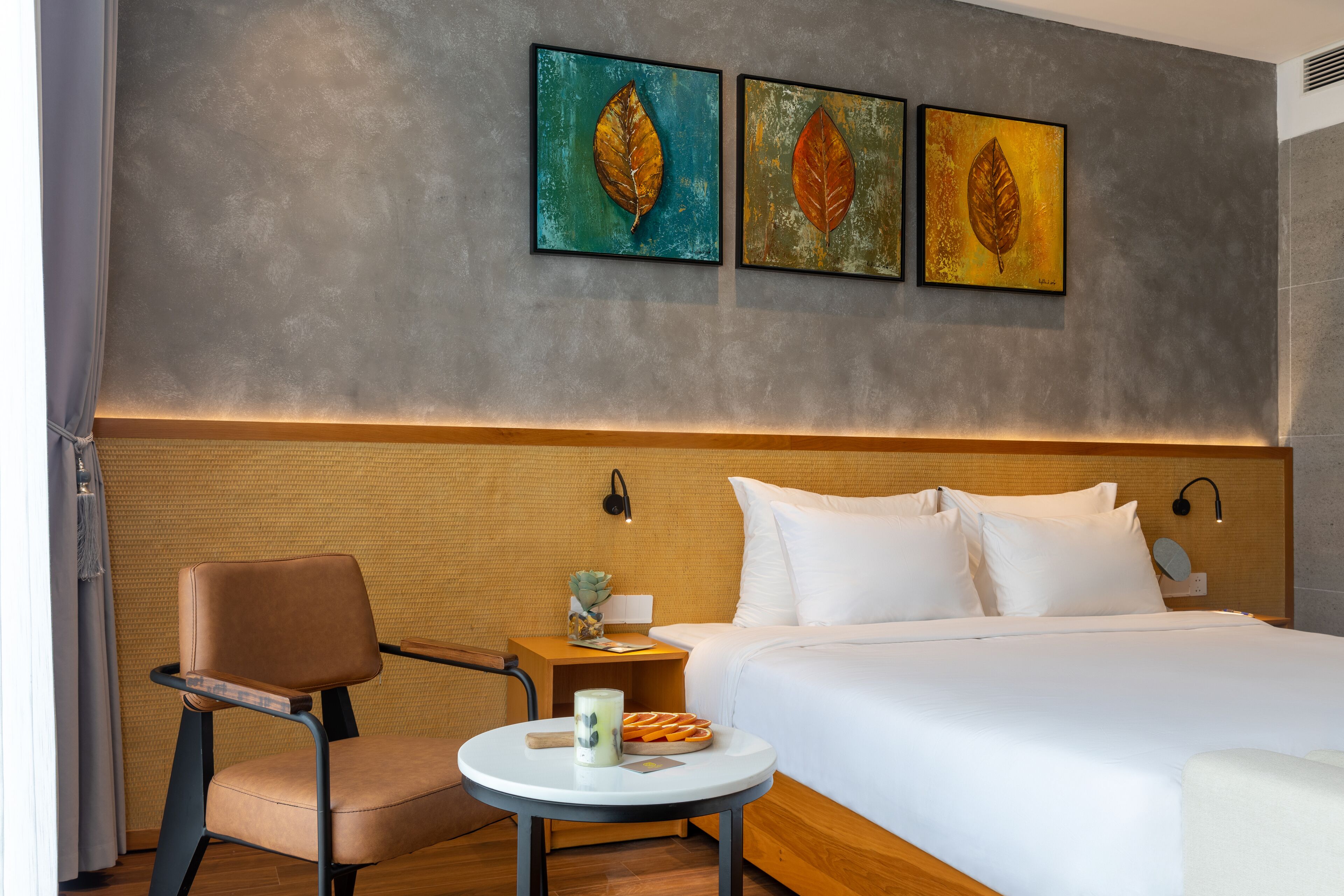 A hotel room with a bed, a chair, a small table with a candle and a plate of fruit, and three leaf paintings on the wall.