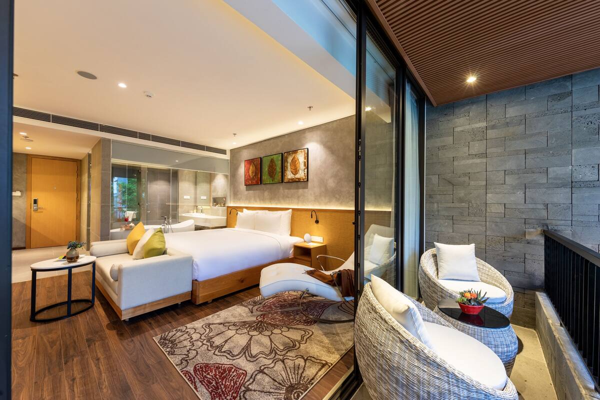 A modern hotel room with a large bed, a sitting area with wicker furniture, a small table with a vase, and a view of the outdoors through a glass wall.