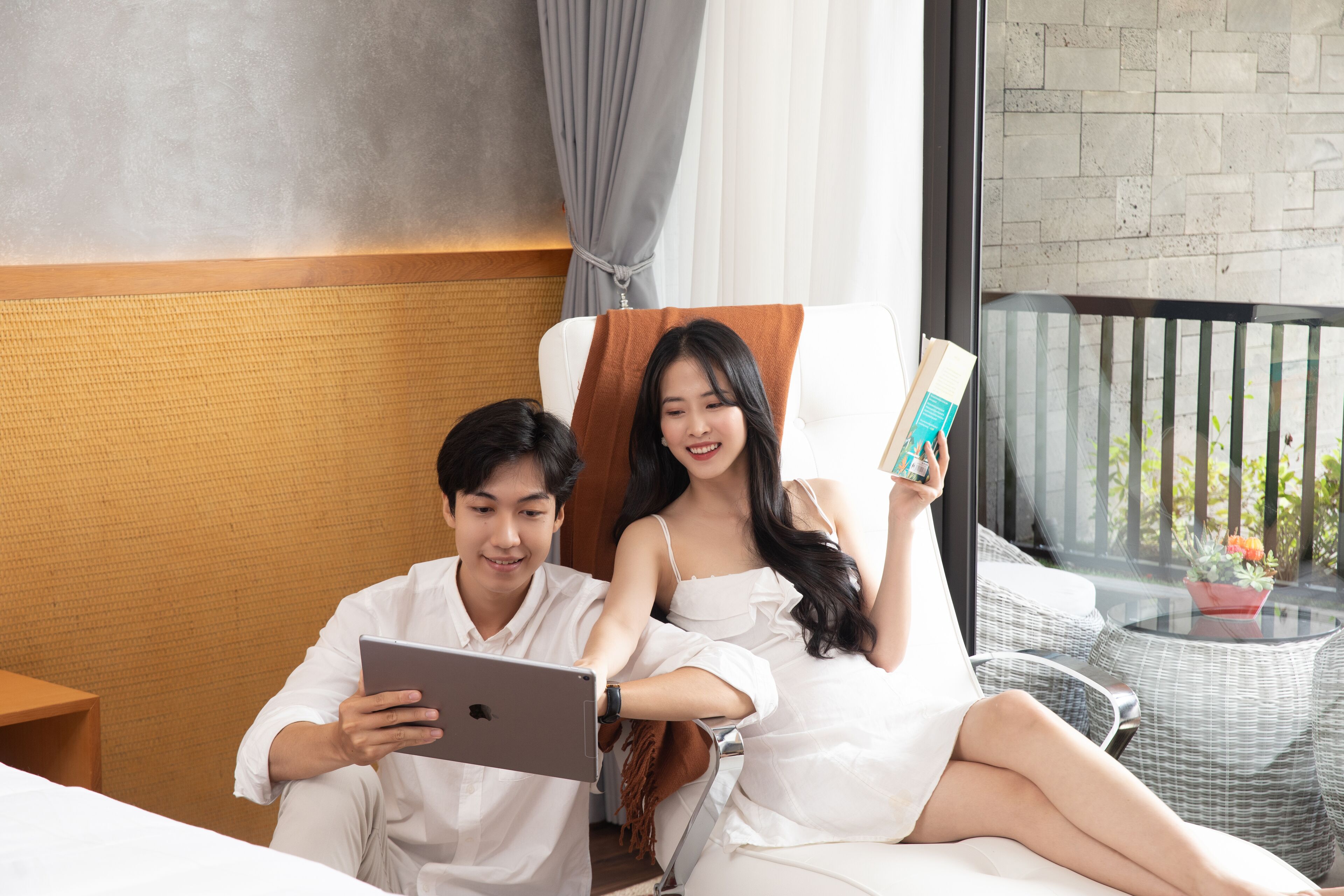A couple sitting on a bed in a hotel room, with a laptop and a book.
