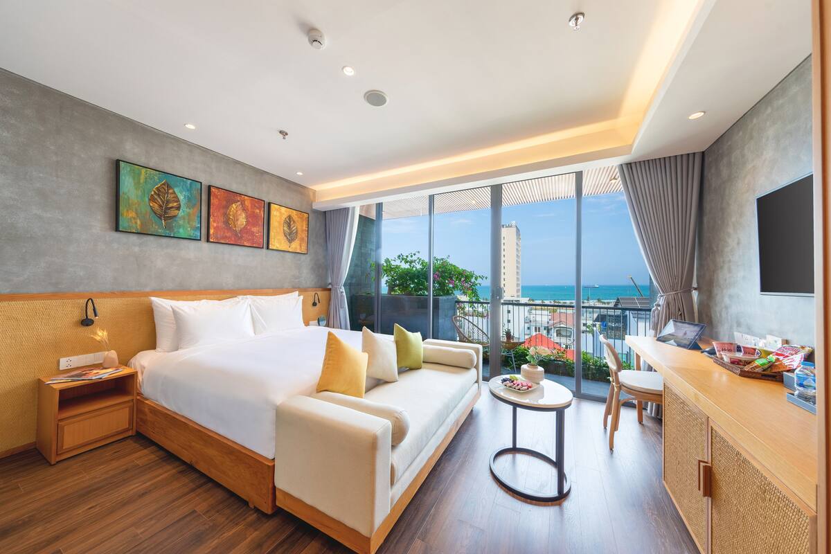 A modern hotel room with a large bed, a sofa, a small table, and a balcony with a view of the city and the sea.