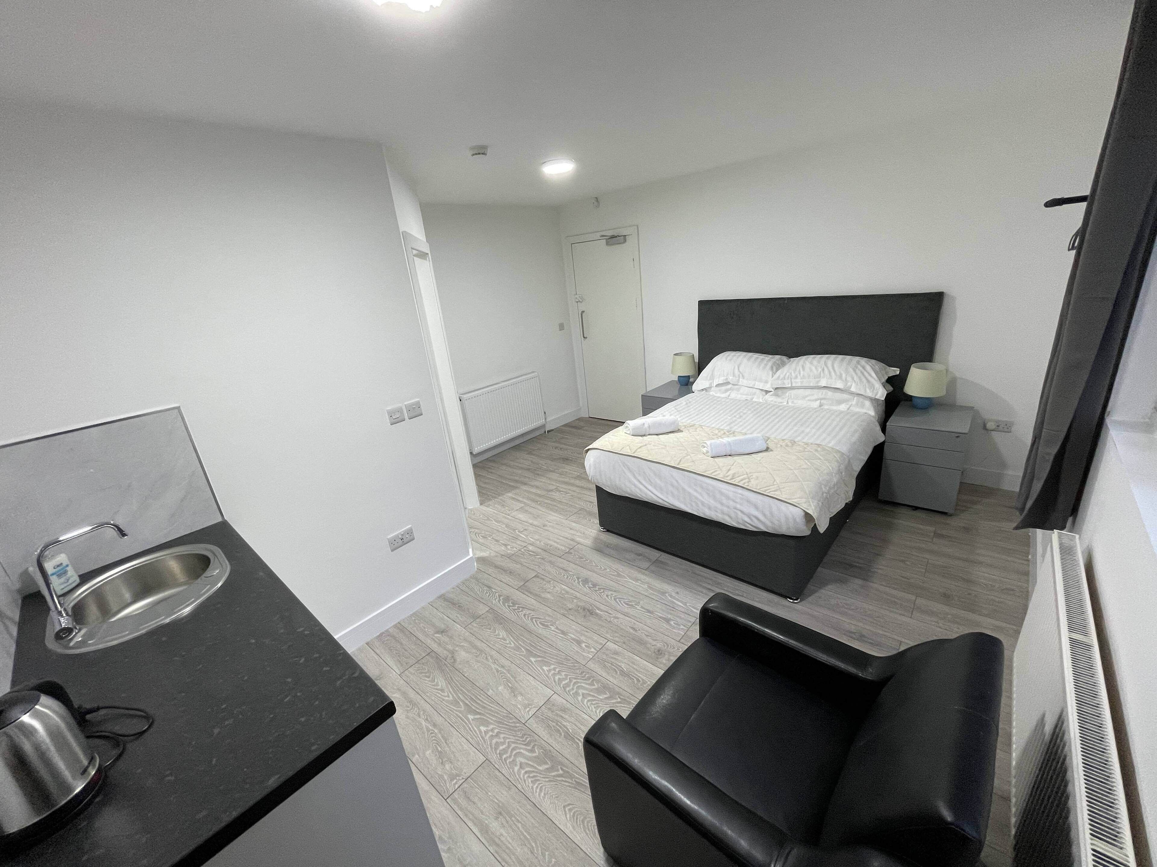 Una habitación de hotel moderna con cama, mesitas de noche, un pequeño escritorio, una silla y una zona de kitchenette.