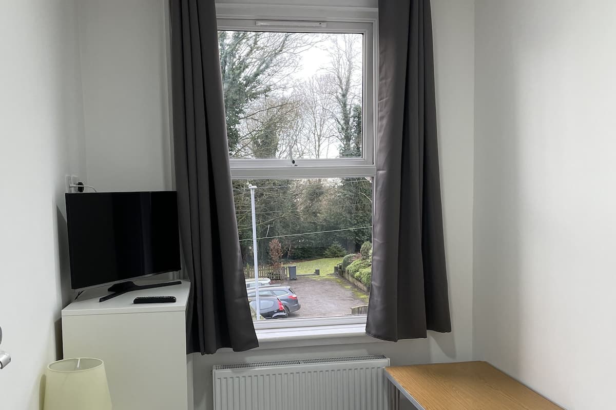 A hotel room with a bed, a desk with a TV, a radiator, a window with curtains, and a view of the outside.