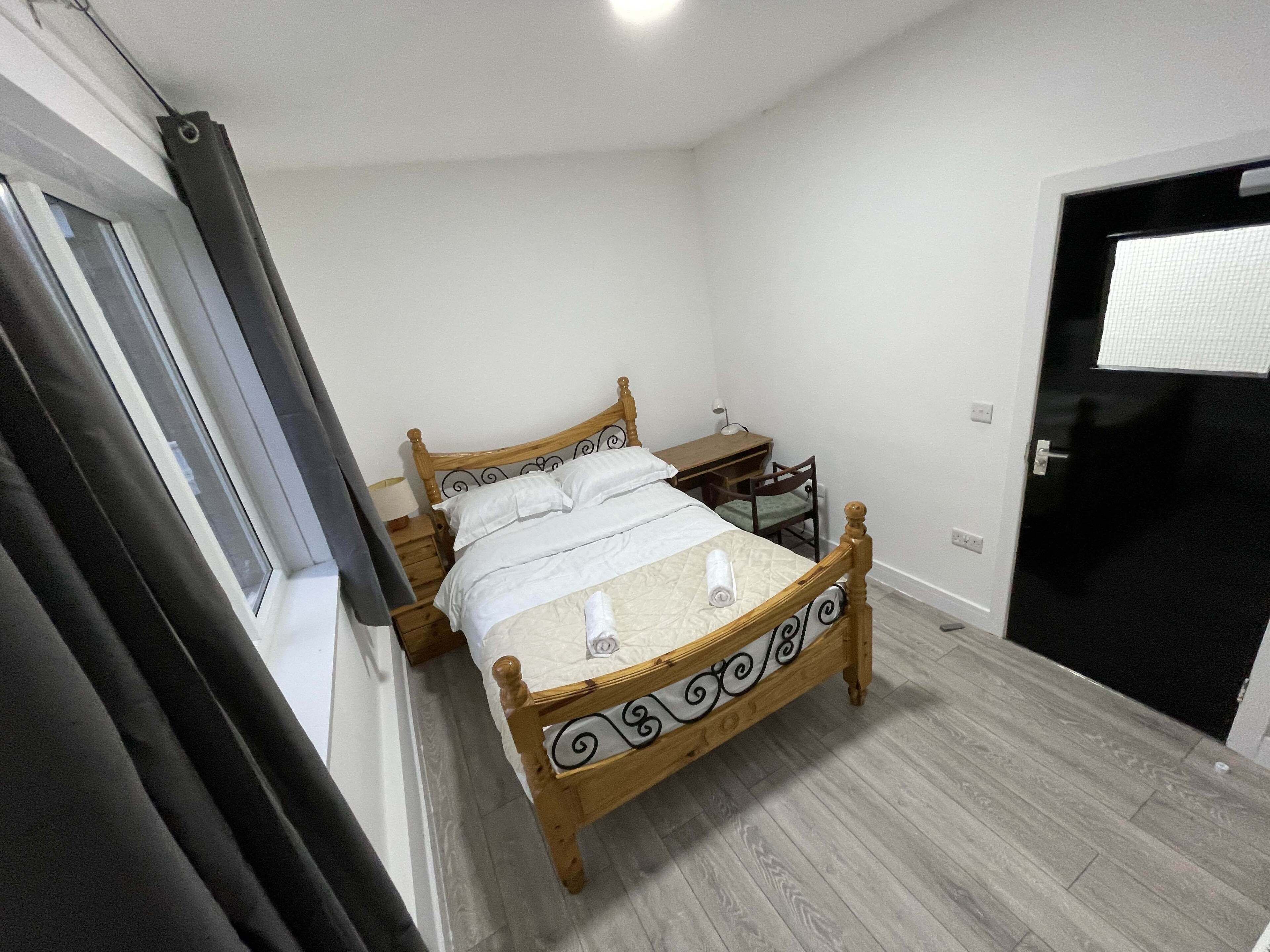 A bedroom with a wooden bed, a bedside table, a chair, a desk, and a window with curtains.