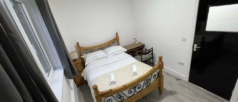 A bedroom with a wooden bed, a bedside table, a chair, a desk, and a window with curtains.