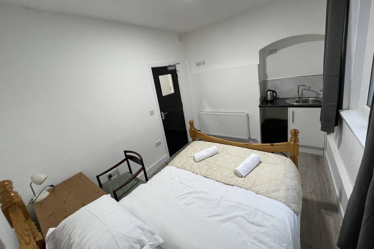 A small, clean room with a bed, a chair, a desk, and a kitchenette.