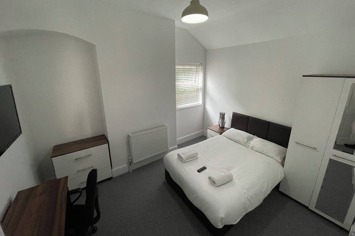 A bedroom with a bed, bedside table, chair, radiator, window with blinds, and a hanging light fixture.