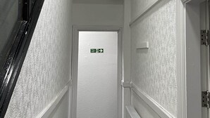 A narrow hallway with textured walls, a visible exit sign, and a radiator on the right. - ISUITES (Newcastle-Under-Lyme)