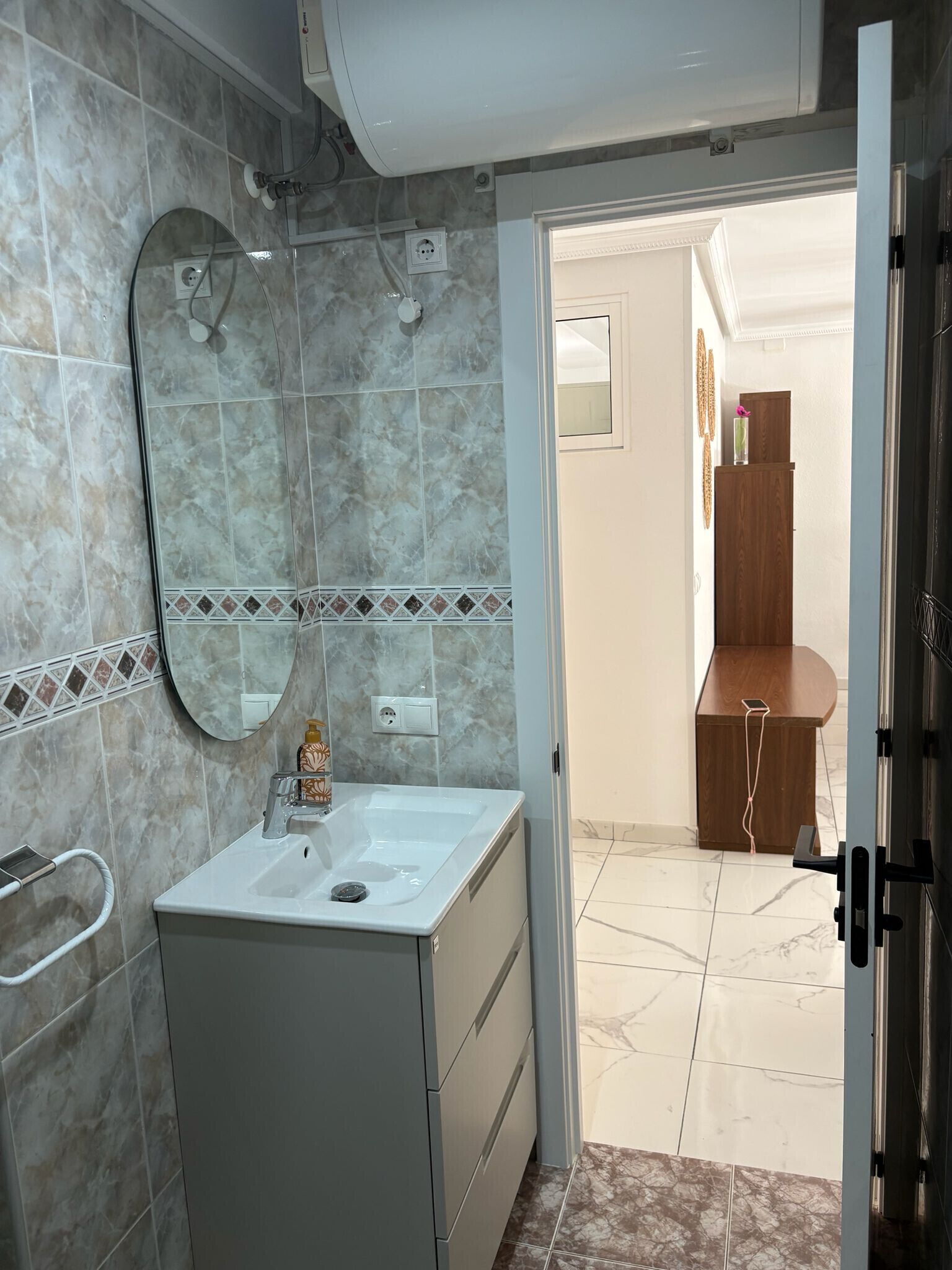 A bathroom with a white sink, mirror, and tiled walls. There is a wooden cabinet and a door leading to another room.