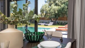 Una mesa de comedor con un jarrón, un cuenco verde y un vaso. Se ve una piscina y follaje a través de la ventana.