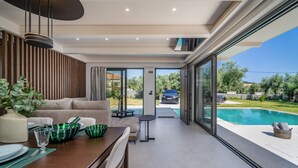 Un comedor moderno con una mesa de madera, cristalería verde y vistas a una piscina y vegetación al exterior.
