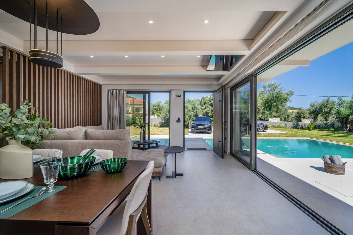 A modern dining area with a wooden table, green glassware, and a view of a swimming pool and greenery outside.