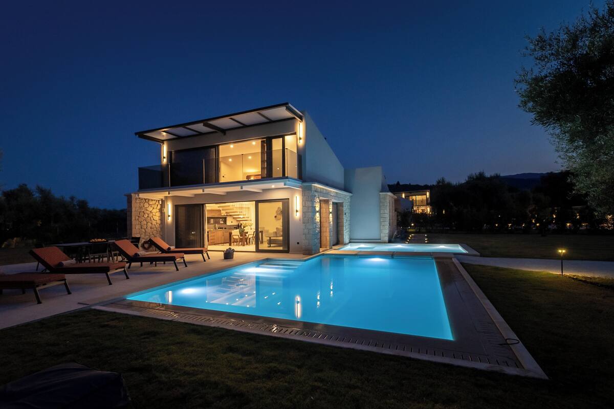 A modern two-story house with a swimming pool, outdoor seating, and a well-lit interior.