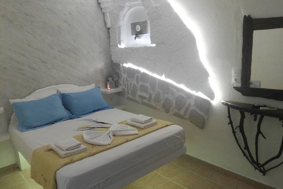 A bedroom with a bed, blue pillows, a bedside table with a lamp, a wall-mounted TV, and a stone wall.