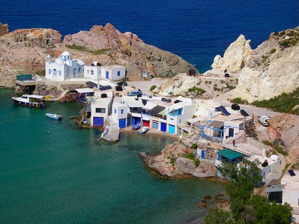 A coastal village with white buildings, a boat docked near the shore, and rocky cliffs in the background.