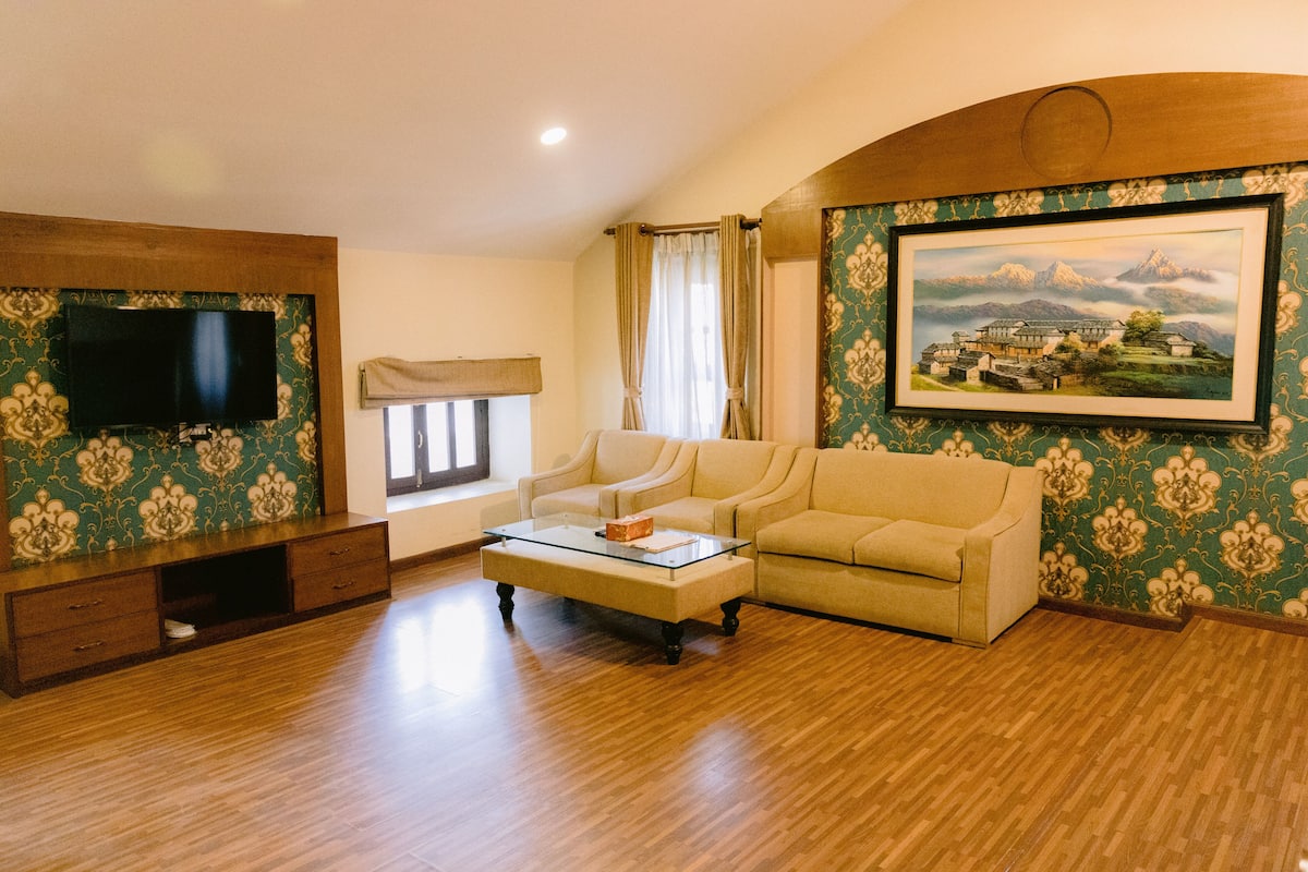 Una sala de estar con un televisor de pantalla plana, un sofá color beige, una mesa de centro de vidrio y un cuadro de un paisaje montañoso.