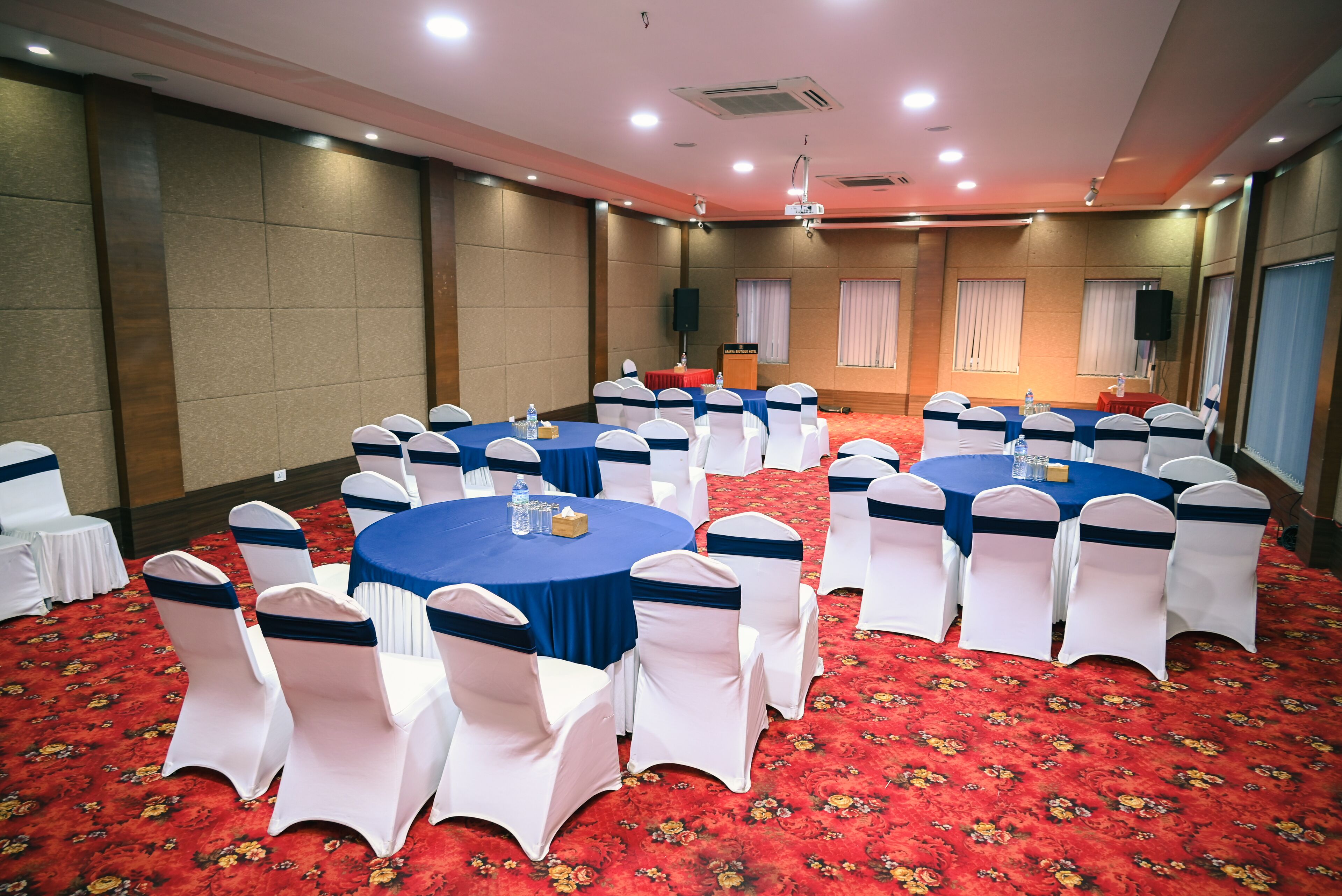 A conference room with round tables, white chairs with blue sashes, a red carpet, and a stage with a podium.
