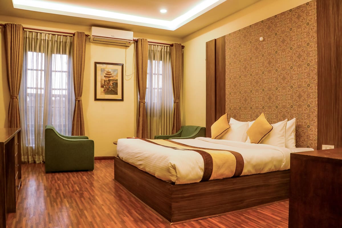 Una habitación de hotel con piso de madera, cama con ropa de cama blanca y detalles marrones, dos sillas verdes y un ventanal con cortinas.