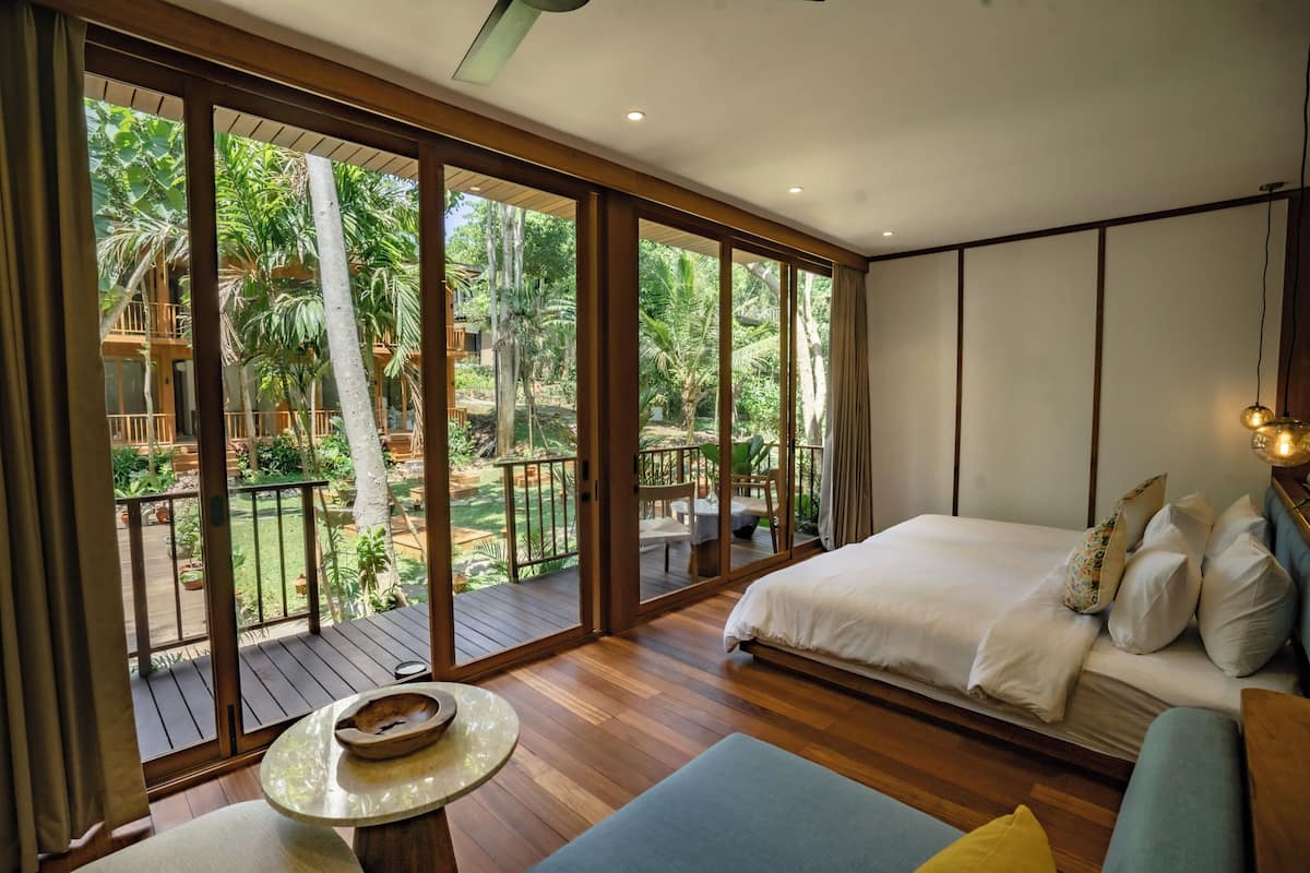 A spacious bedroom with a large bed, a seating area, and a balcony with a view of a lush garden.