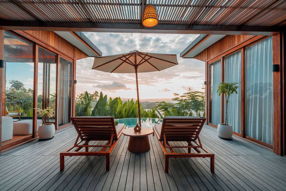 A wooden deck with two wooden chairs and a table, a large umbrella, and a view of a swimming pool and landscape.