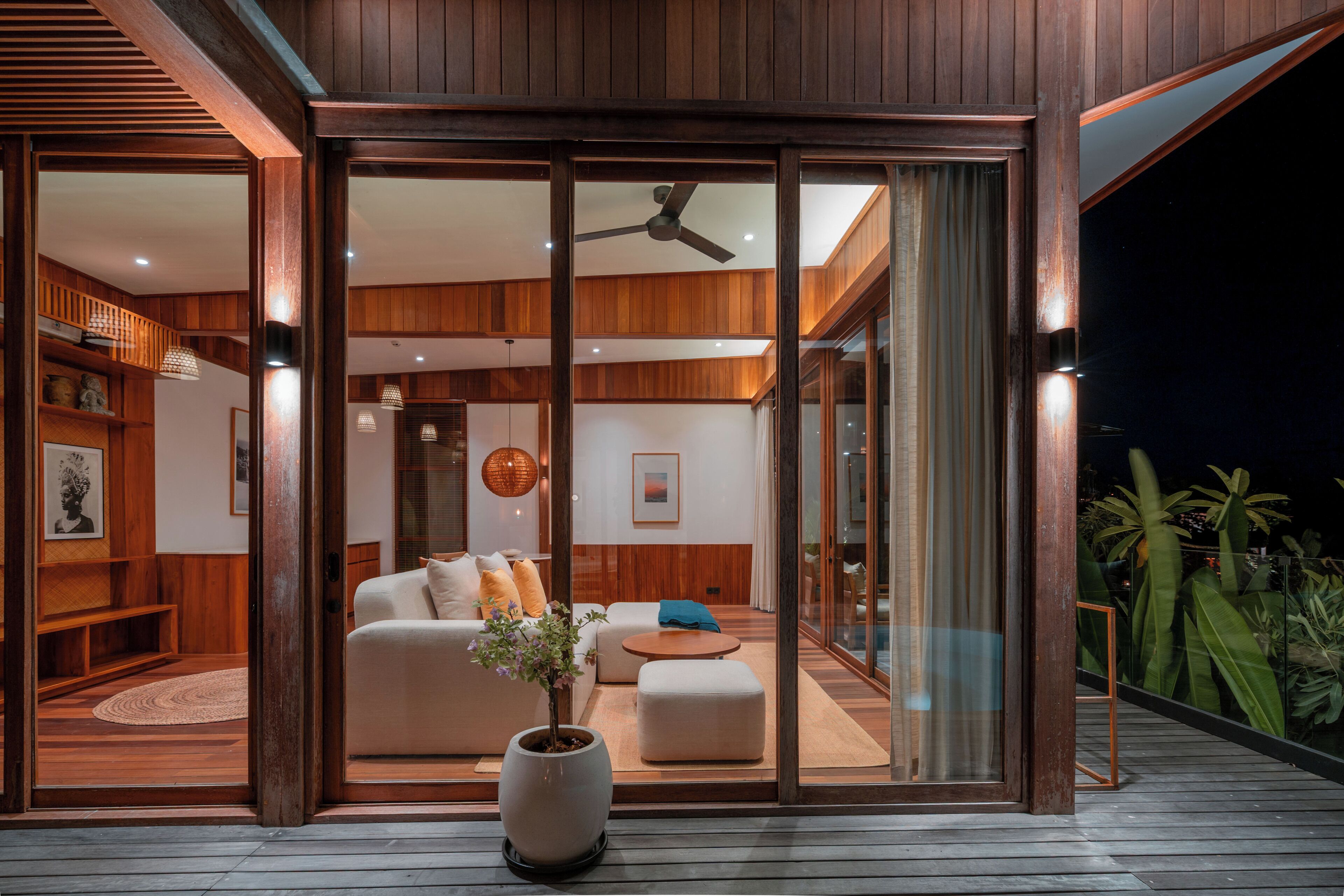A modern interior with a wooden deck, a large sliding glass door leading to a living area with a sofa, ottoman, and a potted plant.