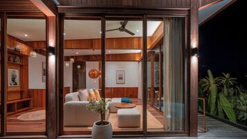 Un intérieur moderne avec une terrasse en bois, une grande porte-fenêtre coulissante donnant sur un espace de vie comprenant un canapé, un pouf et une plante en pot.