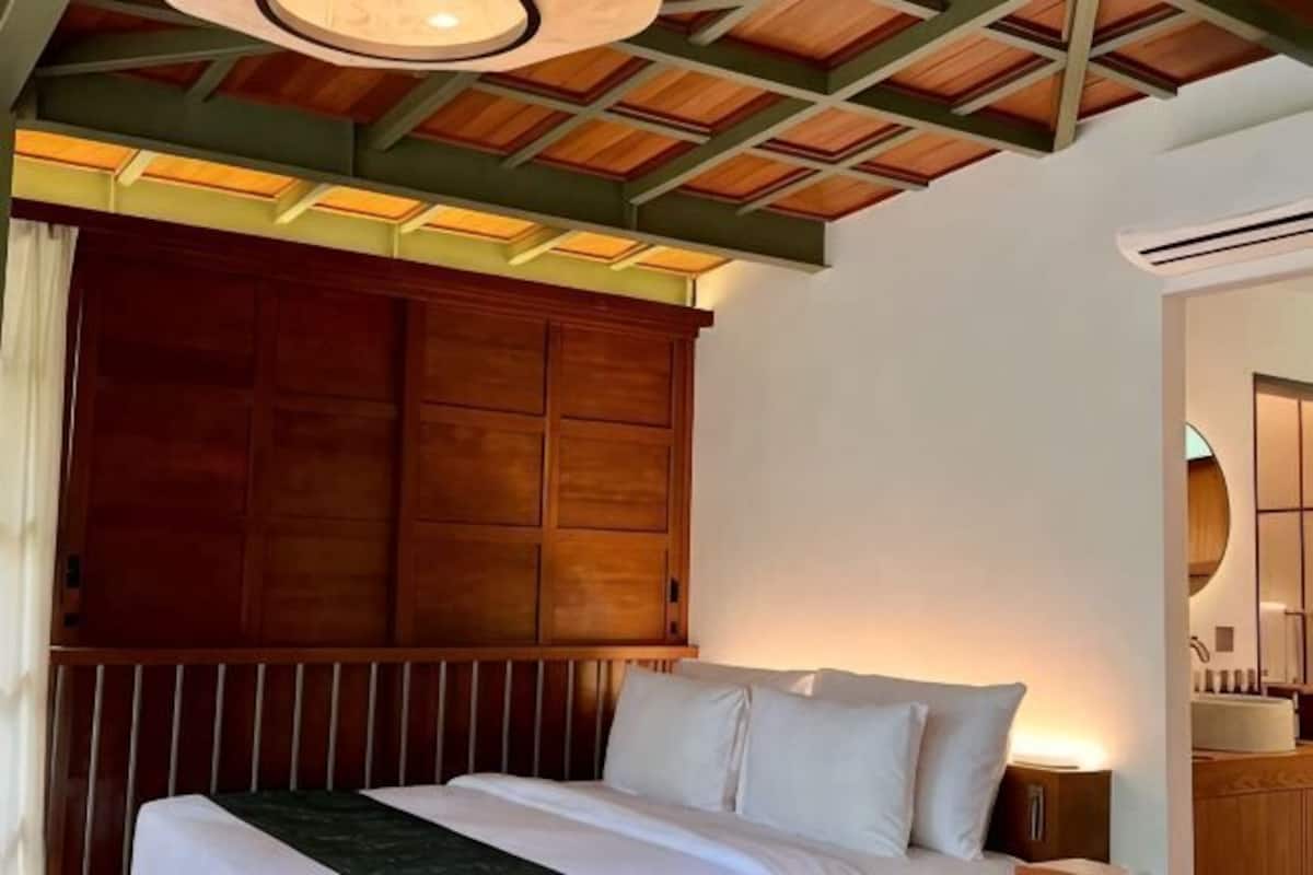 A hotel room with a wooden headboard, a bed with white linens, a nightstand, and a ceiling with wooden beams.