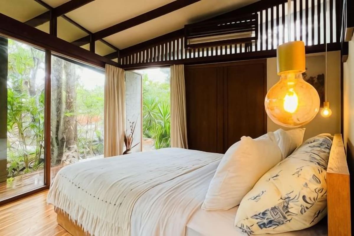 A bedroom with a large bed, a hanging light fixture, and a view of greenery outside.