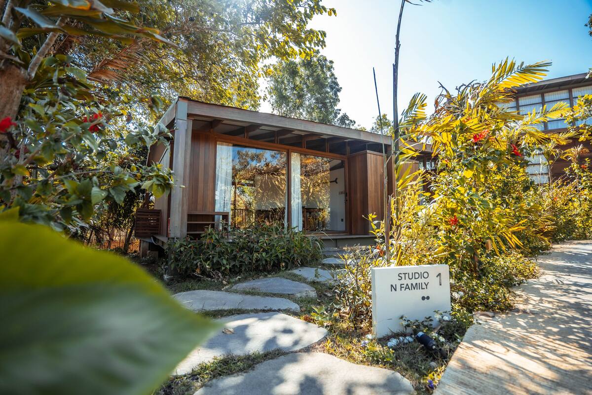 A modern, minimalist studio with a wooden exterior and large windows, surrounded by lush greenery and a stone pathway.