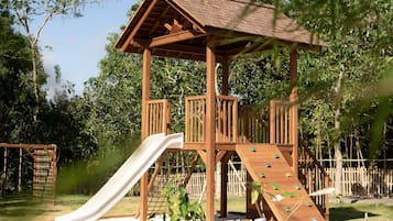 A wooden play structure with a slide and climbing area, surrounded by grass and trees.