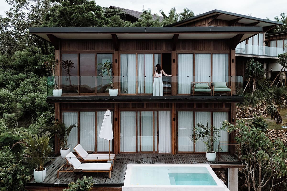 A modern two-story house with a balcony, a pool, and lounge chairs.