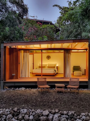 A modern, glass-enclosed bedroom with a bed, two chairs, and a nightstand.
