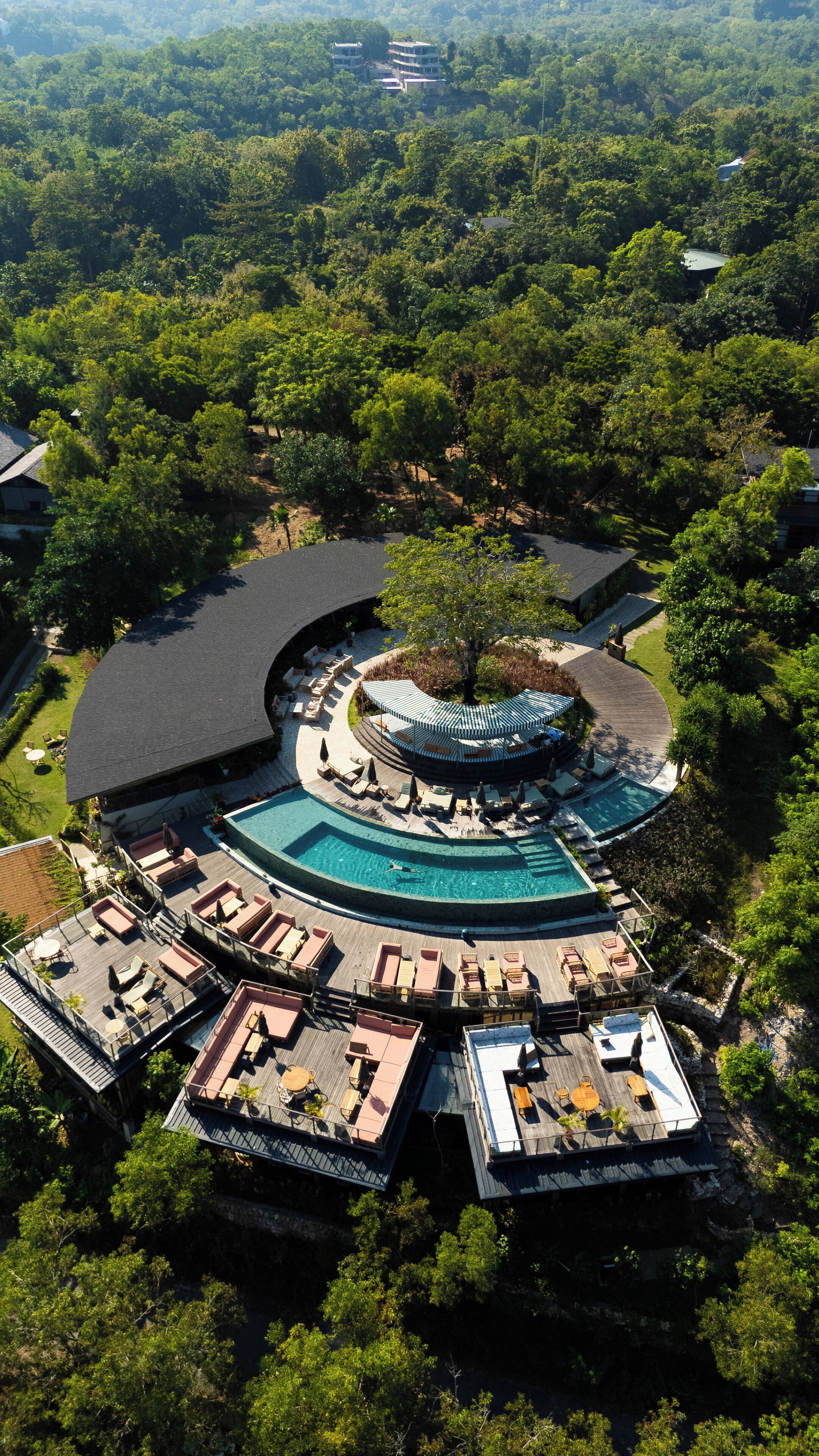 An aerial view of a resort with a central pool, multiple deck areas with seating, and lush surrounding greenery.