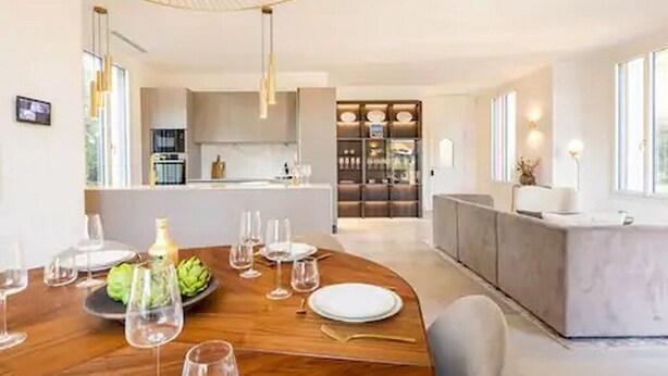Un moderno comedor con una mesa de madera dispuesta para una comida, una cocina al fondo y un amplio ventanal que proporciona luz natural.
