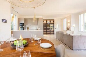 Een moderne eetruimte met een houten tafel gedekt voor een maaltijd, een keuken op de achtergrond en een groot raam dat natuurlijk licht binnenlaat.